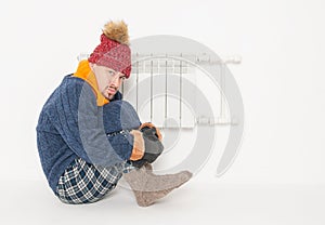 Man feeling cold in hat and pullover sitting close to radiator. Gas crisis concept