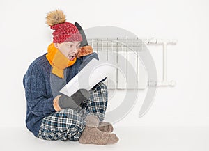 Man feeling cold in hat and pullover sitting close to radiator and hold high electricity gas bill on white background