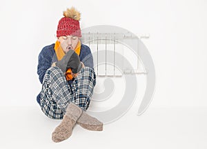 Man feeling cold in hat and pullover sitting close to radiator on white background