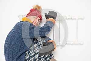 Man feeling cold in hat and pullover sitting close to radiator on white background
