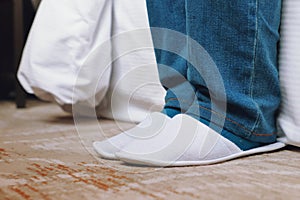 Man feet wearing white hotel slippers, wearing jeans pant and sitting on white bed in hotel