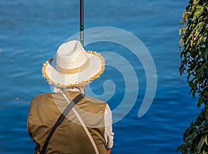 Man fishing with a cane, sitting on rocks with a straw hat on his back.