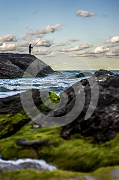 man fishing in the sea in the background and landscape with rocks, seaweed and rain-laden clouds.
