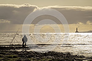 Man fishing silhouetted against a bright golden sea with roker pier and lighthouse blurred in the background.