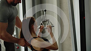 A man fitness instructor in the gym helps a woman use exercise machine and maintains her correct posture. Slender and