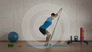 Man fitness instructor in sportswear doing half squat rubber band exercise at the home studio.