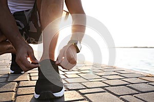 Man with fitness tracker tying shoelaces near river, closeup. Space for text