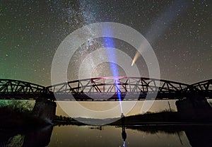 Man with flashlight standing on river bank under illuminated metal bridge under dark starry sky and Neowise comet with light tail