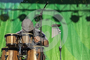 Man with flat cap playing wooden Caribbean bongo drums with mircophone and chimes and green curtain behind