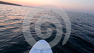 Man floating white SUP board paddles rowing oar POV dawn sunset sea surface wave