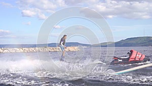 Man is flying on flyboard on lake or sea in day, hydrocycle is floating near, water flows is lifting human