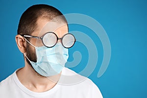 Man with foggy glasses caused by wearing disposable mask on blue background, space for text. Protective measure during coronavirus