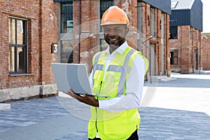 Man foreman in reflective vest and white helmet using portable laptop at construction site. Concentrated african