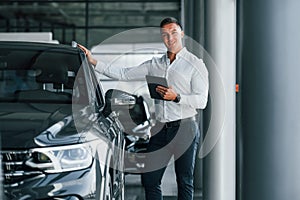 Man in formal clothes is standing indoors near moden new car