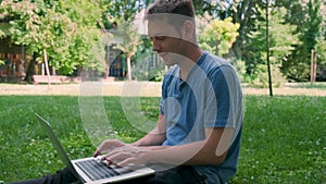 Man freelancer in blue t-shirt sit on green grass in city park, work with laptop and drinking coffee from paper cup