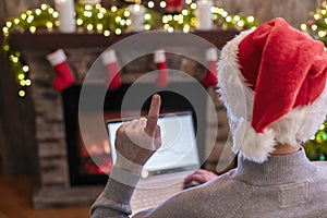 Man freelancer in santa claus hat working on laptop having idea and raising a finger up sitting near christmas tree and fireplace.