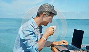Man freelancer working at morning on the beach by the sea, drinking coffee, using laptop computer.