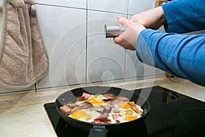 The man fries bacon and sausage in a pan and adds eggs and salt to the eggs