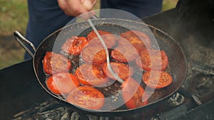 Man is frying a slices of tomates on frying pan with oil on coals in the grill.