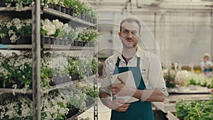 Man gardener is examining flowers holding the digital tablet and looking at the camera. Male greenhouse worker portrait