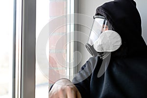 A man in a gas mask and a black hood looks out the window sitting against the wall