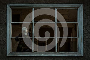 A man in a gas mask looks out of a window without glass, standing in a ruined house