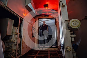 Man in gateway corridor of airlock of abandoned bunker. Big hermetic door illuminated by red lantern