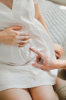 A man gently touches his wife& x27;s pregnant belly. Close-up of the palms, stomach. White background and light clothes.