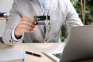 Man with gift card and laptop at table indoors, closeup