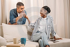 african american man gifting ring in heart shaped box to woman sitting on couch