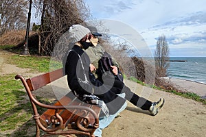 A man and a girl in medical masks sit on a park bench and look at the sea horizon