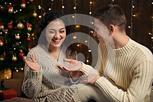 The man gives the girl an engagement ring, couple is in christmas lights and decoration, dressed in white, fir tree on dark wooden