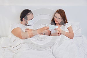 A man gives a woman a medical mask while lying in a white bed, concept. Relationship problems coupled with isolation due to