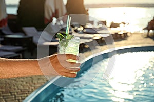 Man with glass of fresh summer cocktail near swimming pool outdoors at sunset. Space for text