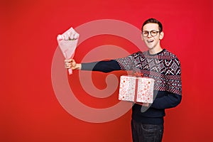 A man in glasses holds a gift and holds out a bouquet of flowers on a red background