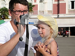 A man in glasses and a little girl in a yellow hat are videotaping. Sunny hot summer day