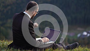 A man in glasses and a suit sits on a hill holding laptop on his lap