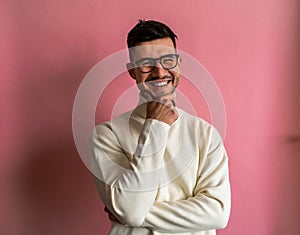A man with glasses and a white sweater stands in front of a pink wall, smiling