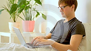 A man with glasses works in the bedroom at a laptop.