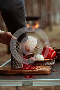 man grates beets and other vegetables outside the house. Cooking outdoors. Close-up. Barbecue in the background