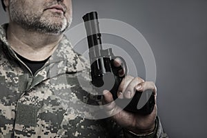 A man with a gray beard and in camouflage blows on the barrel of a pistol. Military conflicts, murders and crimes. Grey background