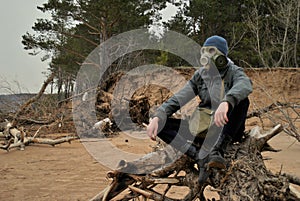 A man in a gray jacket, dark jeans and a gas mask sits in the spring evening on the banks of the Volga River on an old dry tree