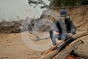 A man in a gray jacket, knitted hat, dark jeans and a gas mask sits on a burning fire in the spring evening and warms his hands.