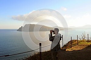 Man in gray military jacket looks through binoculars on high altitude mountain surrounded by beautiful sea and mountains in the