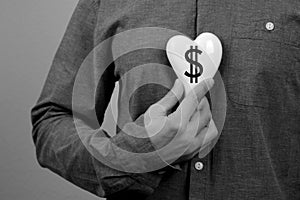 Man in a gray shirt in his hand near his chest holds a white heart with a dollar symbol, the concept of commercialism, self- photo