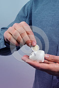 Man in a gray shirt puts a 2 euro metal coin into a small white piggy bank, a concept of saving and saving money, banking