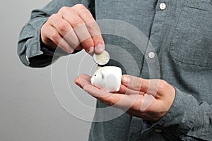 Man in a gray shirt puts a 2 euro metal coin into a small white piggy bank, a concept of saving and saving money, banking