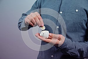 Man in a gray shirt puts a 2 euro metal coin into a small white piggy bank, a concept of saving and saving money, banking