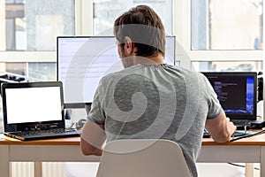 A man in a gray t-shirt is sitting at a workplace with two laptops and a monitor near the window. Remote work from home