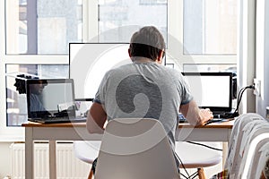 A man in a gray t-shirt is sitting at a workplace with two laptops and a monitor near the window. Remote work from home